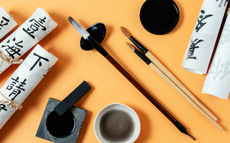A flat lay of traditional calligraphy tools on an orange background, including rolled sheets of paper with Chinese characters, ink brushes of various sizes, an ink stone, and small bowls of black