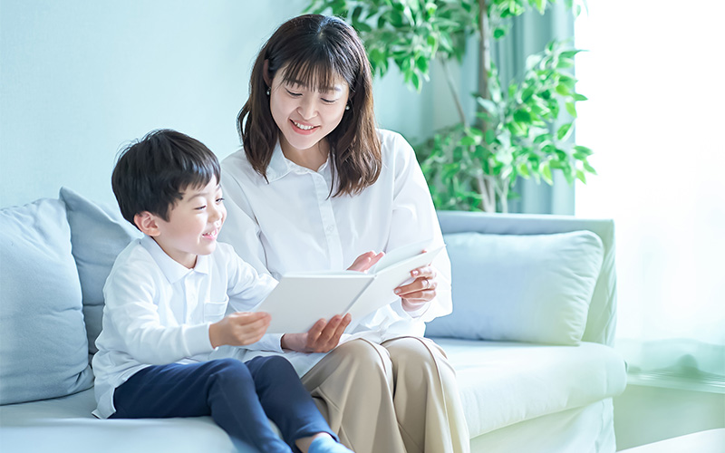 A mother and young son sitting on a light-colored sofa, smiling while reading from a tablet together in a bright living room with green plants in the background