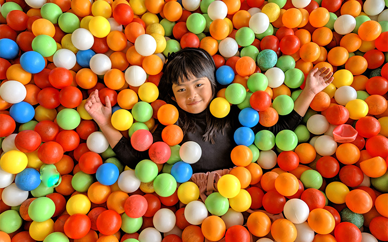 A young child smiling and lying on their back in a colorful ball pit, surrounded by bright red, yellow, green, blue, and white plastic balls with arms stretched out
