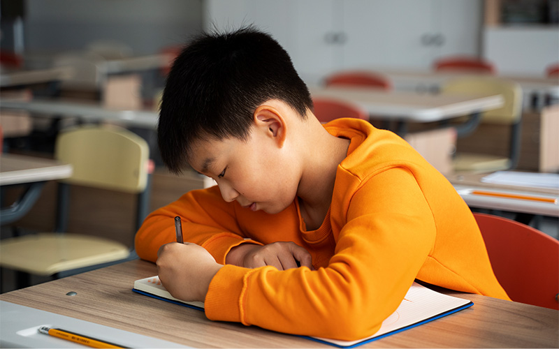A young student wearing an orange sweatshirt sits at a classroom desk, leaning forward and writing in a notebook, with empty desks and chairs visible in the background