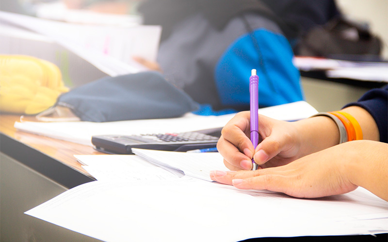 A close-up of a student's hand using a purple pen to write on a paper at a desk, with a calculator and other students blurred in the background.