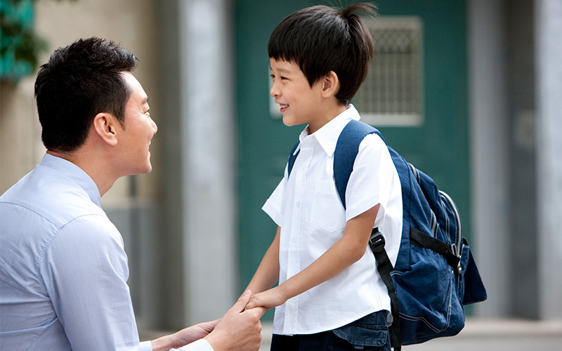 A father crouches down to hold his young son's hand as the boy, wearing a backpack, smiles and gets ready for school.