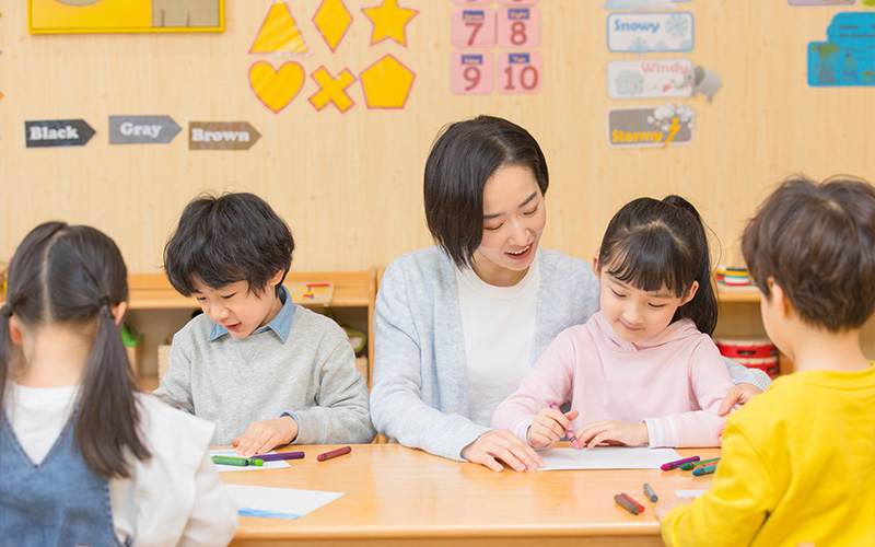 A Chinese language teacher smiling while guiding young students during a preschool drawing activity.
