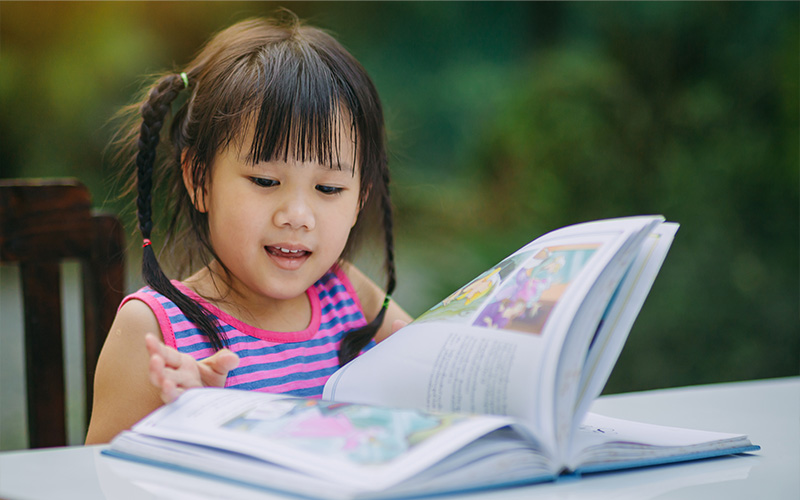 A young girl focused on reading a Chinese picture book outdoors.