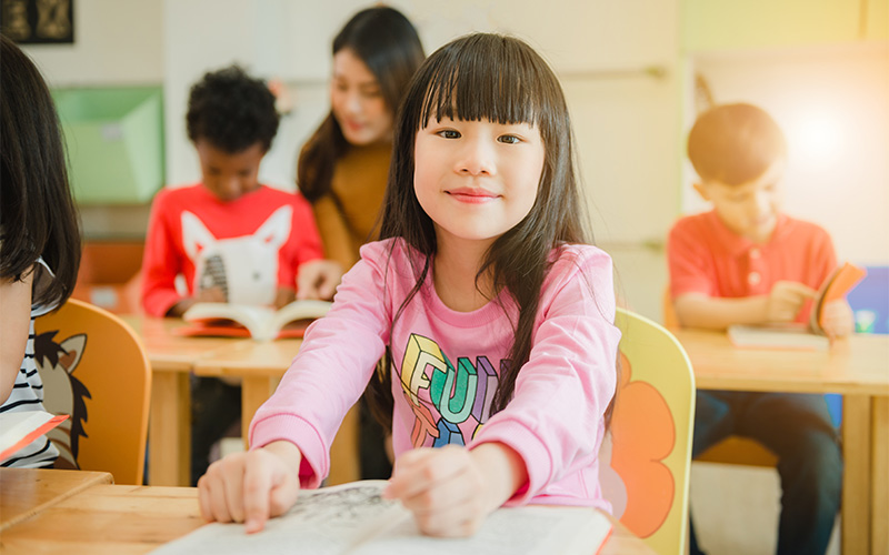 Smiling girl reads book in bright classroom with other children.
