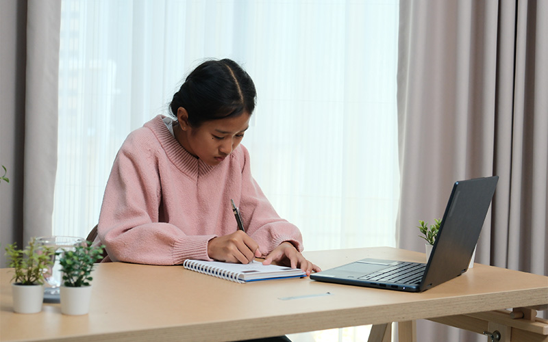 student writing Chinese composition with laptop and notebook.